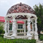 full view of an 8-column round garden gazebo, crafted from white marble with hand-carved female caryatid statues as columns, topped with an ornate copper dome for luxury outdoor use
