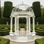 Full view of round white marble column gazebo with white iron dome, set in a formal hedge maze garden