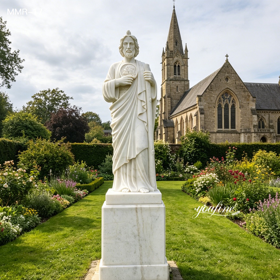 White marble St. Jude statue with a square pedestal, standing in a well-maintained garden beside a historic church.