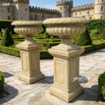 Pair of beige marble planters with ribbed bowls and carved pedestals, set in a formal garden.