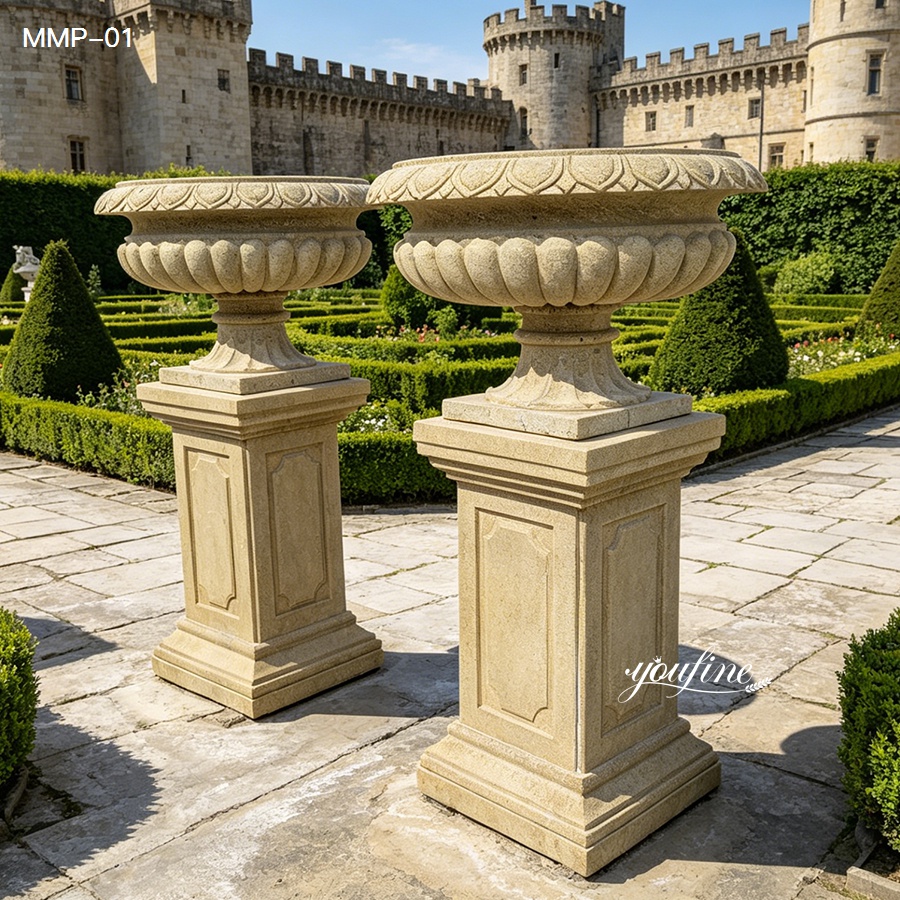 Pair of beige marble planters with ribbed bowls and carved pedestals, set in a formal garden.