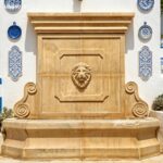 Artisan-carved beige marble wall water fountain set against a wall with blue ceramic tiles