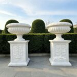 Pair of white marble planters with ribbed bowls, set on a paved patio with trimmed hedges.