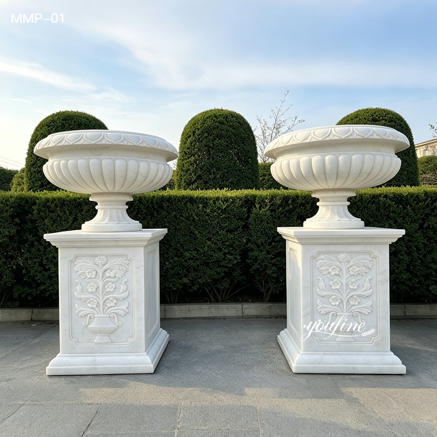 Pair of white marble planters with ribbed bowls, set on a paved patio with trimmed hedges.