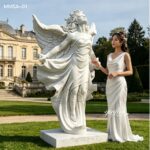 Woman in a white dress standing next to a large marble angel statue, highlighting its elegant design and scale