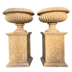 A pair of intricately carved marble urns resting on ornate pedestals, each adorned with floral motifs, against a white background.