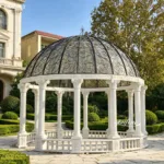 White marble gazebo featuring intricate craftsmanship, decorated with a wrought iron dome, surrounded by manicured greenery.