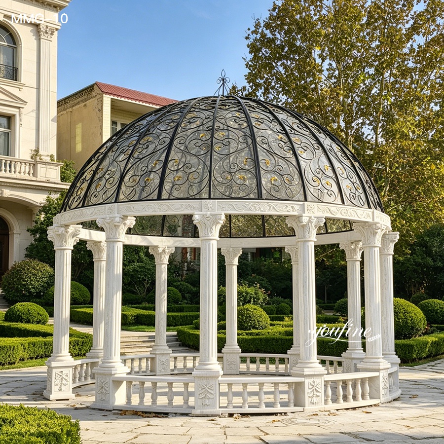 White marble gazebo featuring intricate craftsmanship, decorated with a wrought iron dome, surrounded by manicured greenery.