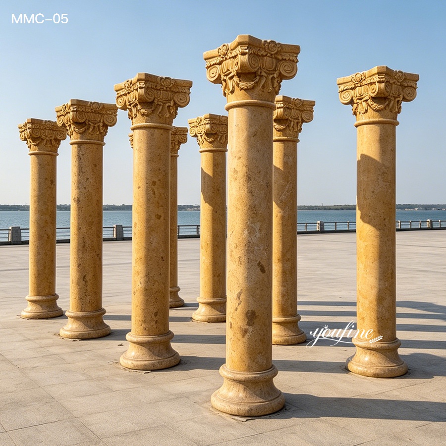 Collection of intricately carved golden marble columns arranged outdoors with a waterfront background under clear blue skies.