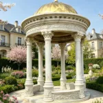 Luxurious marble gazebo with carved columns and a golden dome, set in a lush garden with blooming flowers and elegant architecture.