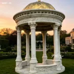 Elegant white marble gazebo with intricate carvings and a golden dome, set in a manicured garden during sunset.