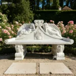 White marble garden bench with intricate angel carvings surrounded by blooming roses in a formal garden setting.
