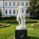 Elegant white marble statue of Bacchus holding a cup, set on a dark pedestal in a lush, flowered garden with a classical building.