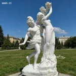 A detailed white marble sculpture of two figures in a classical style, displayed outdoors on a sunny day with a park setting.