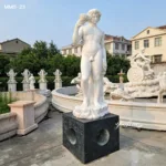 A detailed marble figure sculpture held on a black base, surrounded by fountain decor in an outdoor setting with trees and buildings.