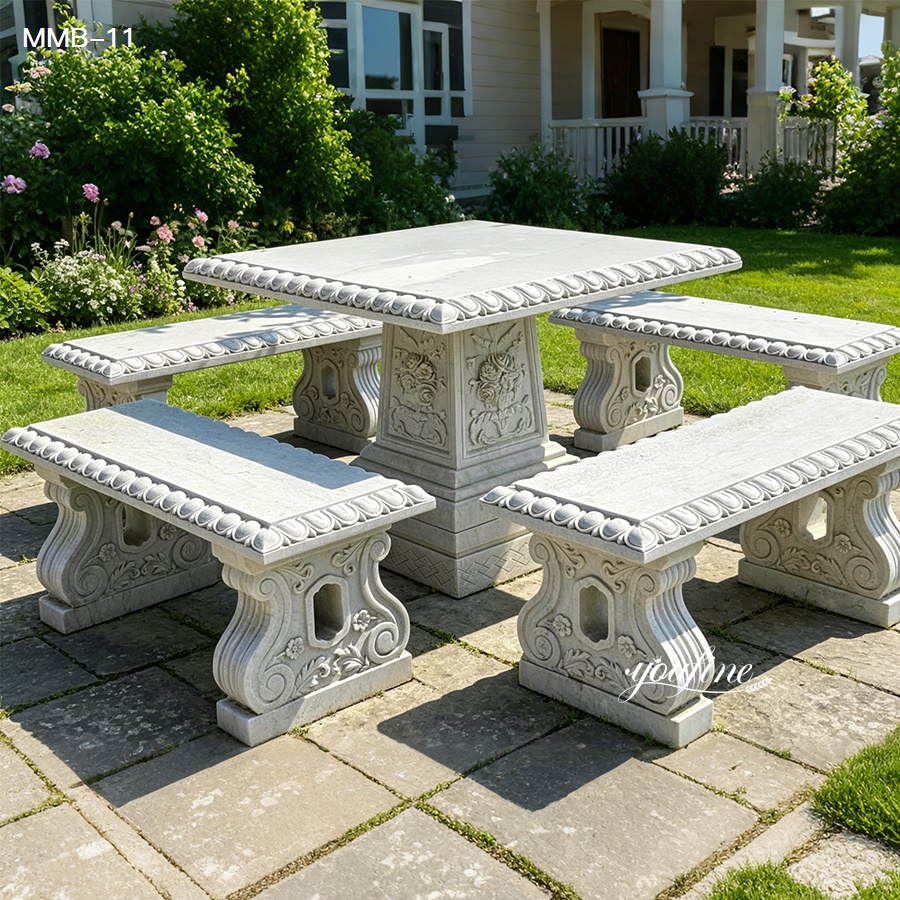 An elegant outdoor marble dining set featuring a rectangular table with intricate floral carvings and four matching benches in a lush garden setting.