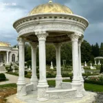 Ornate marble gazebo with a golden dome and Corinthian columns, set in a lush formal garden with pathways and decorative landscaping.