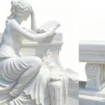 A finely carved white marble sculpture of a woman reading a book, featuring flowing drapery and intricate details on a decorative bench.
