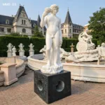 Elegant marble statue of a boy holding a cup, displayed on a black pedestal in a garden with ornate sculptures and a grand architectural backdrop.
