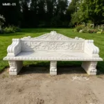 Elegant white marble bench with intricate carvings placed on a manicured garden lawn with trees and flowers.