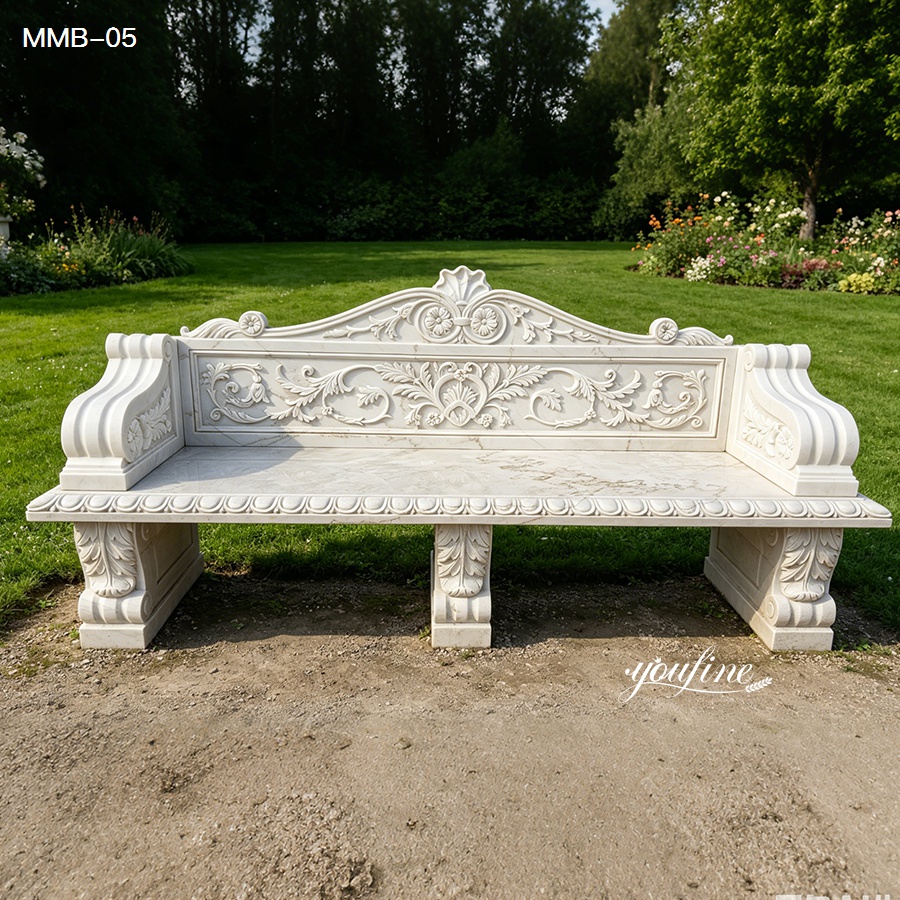 Elegant white marble bench with intricate carvings placed on a manicured garden lawn with trees and flowers.
