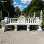 White marble bench with intricate design, featuring lion sculptures on sides, in a landscaped garden with statues and greenery.