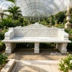 Intricately carved white marble bench with floral details placed within an elegant greenhouse surrounded by lush greenery and blooms.