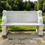 Elegant outdoor marble bench with intricate carvings and decorative motifs placed on a garden path surrounded by green grass.