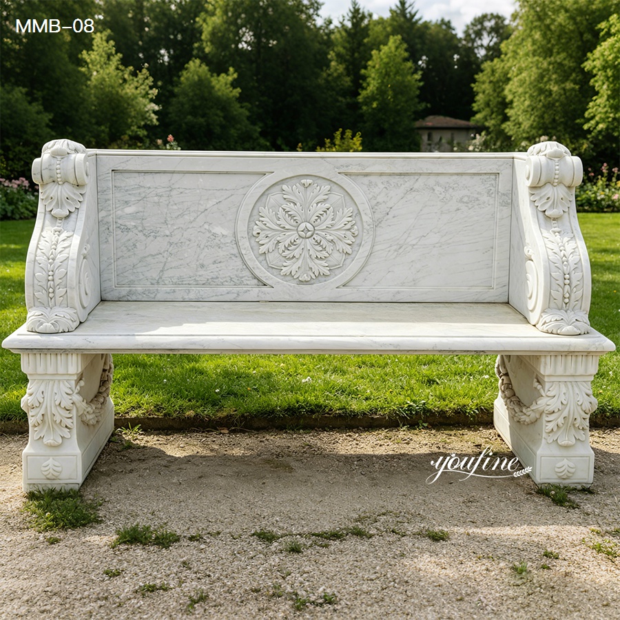 Elegant outdoor marble bench with intricate carvings and decorative motifs placed on a garden path surrounded by green grass.