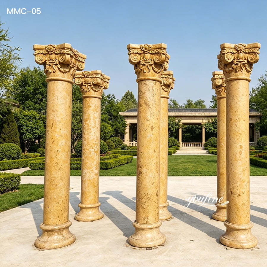 Six ornate marble columns with intricate capitals standing on a paved surface in a meticulously landscaped garden.