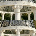Close-up view of a marble gazebo showcasing intricately carved floral columns and decorative wrought iron dome in a garden.