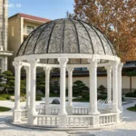 Intricately carved white marble gazebo with a decorative iron dome, surrounded by a landscaped garden with neatly trimmed greenery.