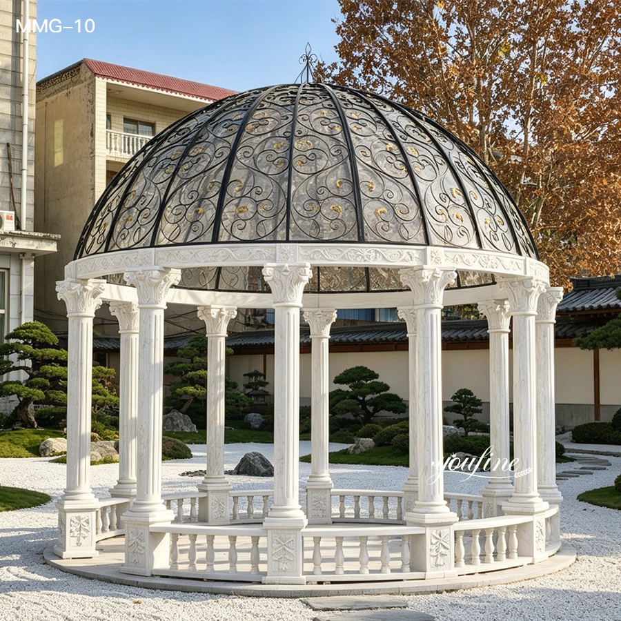 Intricately carved white marble gazebo with a decorative iron dome, surrounded by a landscaped garden with neatly trimmed greenery.