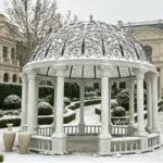 An ornate white marble gazebo with intricate carvings and a decorative iron dome in a snowy landscaped garden.