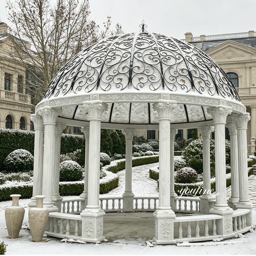 An ornate white marble gazebo with intricate carvings and a decorative iron dome in a snowy landscaped garden.