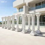 A row of intricately designed white marble columns with ornate details displayed in front of a modern villa featuring glass balconies and clean architecture.