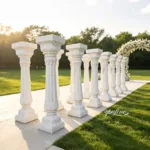 Elegant white marble columns with intricate carvings arranged outdoors on a sunny day, with a floral arch in the background.