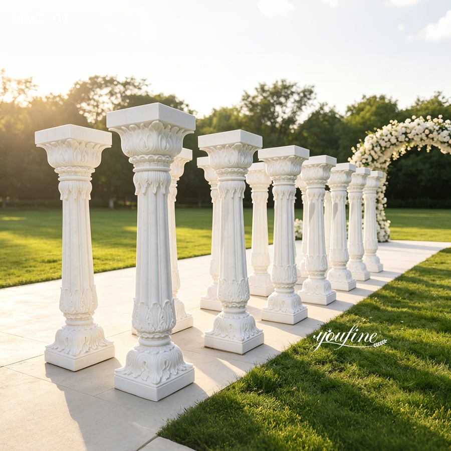 Elegant white marble columns with intricate carvings arranged outdoors on a sunny day, with a floral arch in the background.