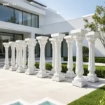 Elegant white marble columns with intricate carvings arranged outdoors near a modern building with landscaped greenery and clean design.