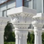 Elegant white marble Corinthian columns featuring intricate floral carvings, displayed outdoors with architectural background.
