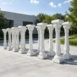 A row of intricately carved white marble decorative columns placed outside in a modern plaza with trees and a building in the background.