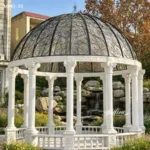 A white marble gazebo with intricate columns and an ornate black iron dome, surrounded by lush foliage and rocks.