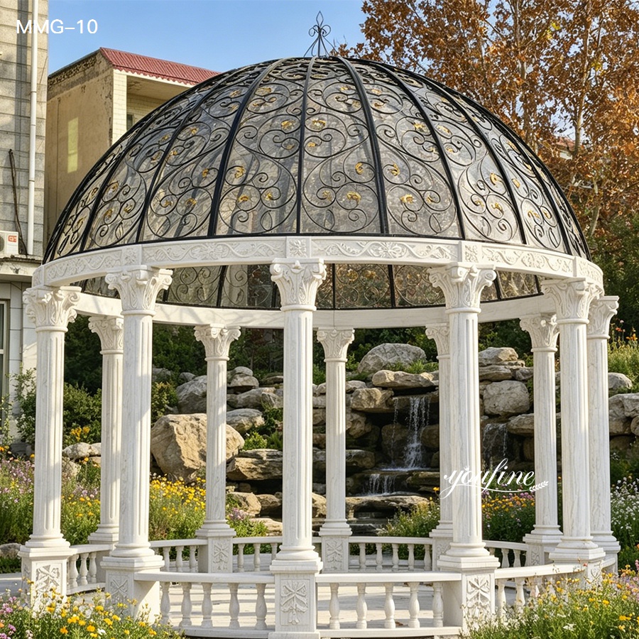 A white marble gazebo with intricate columns and an ornate black iron dome, surrounded by lush foliage and rocks.