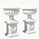 Two elegant white marble urns mounted on pedestal bases with intricate decorative carvings, showcasing classic luxury design.