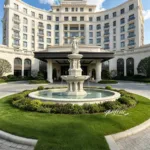 A grand marble fountain with angel sculptures at the entrance of a luxury hotel surrounded by manicured landscaping and greenery.