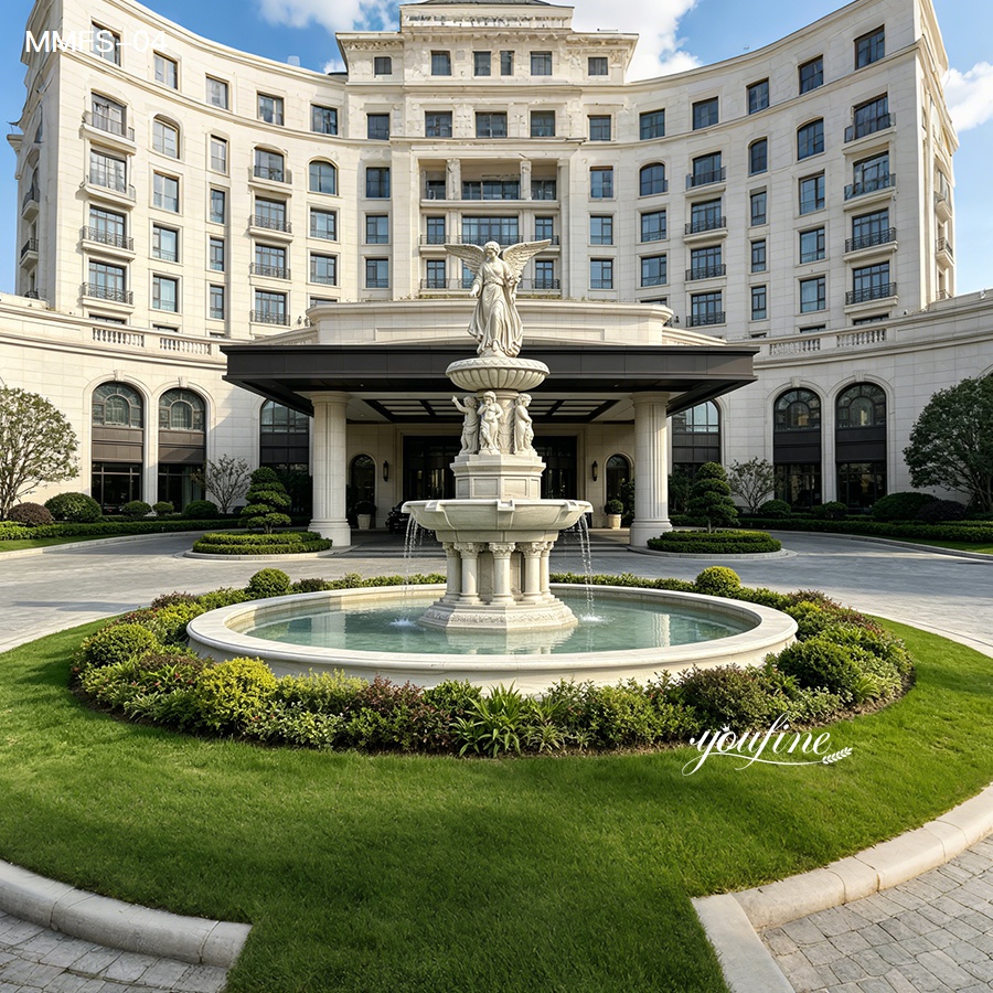A grand marble fountain with angel sculptures at the entrance of a luxury hotel surrounded by manicured landscaping and greenery.