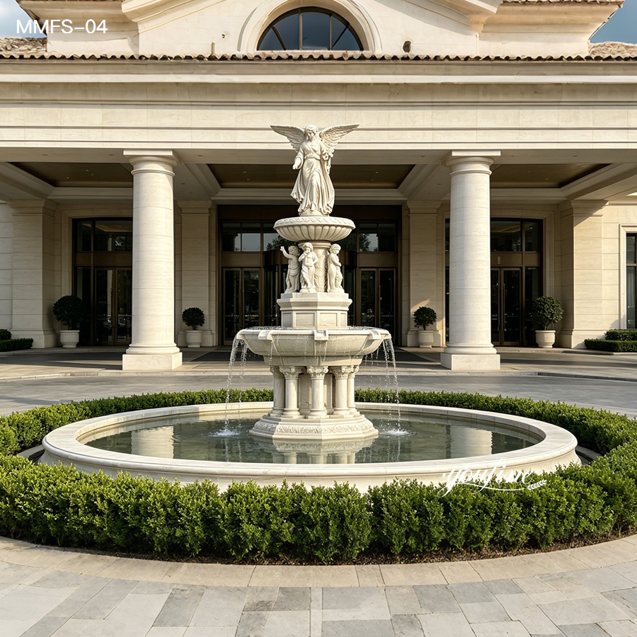 Ornate marble fountain with angel statue centerpiece, surrounded by trimmed greenery and located in front of a grand building facade with columns.