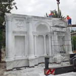 Large white marble wall sculpture with intricate carvings and columns being installed by workers on scaffolding outdoors.