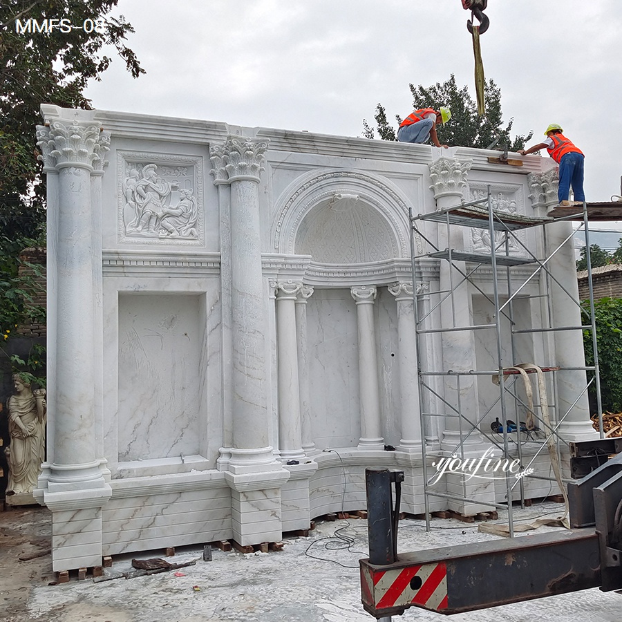 Large white marble wall sculpture with intricate carvings and columns being installed by workers on scaffolding outdoors.