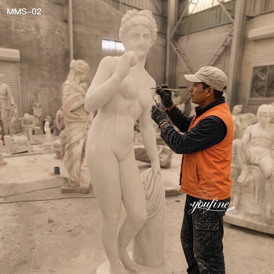 An artisan wearing an orange vest carefully carving a marble statue of a graceful female figure in a workshop filled with sculptures.
